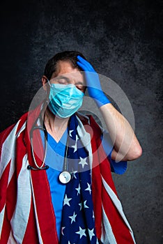Stressed doctor with hand on head, wearing face mask and holding the American flag