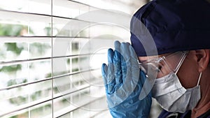 Stressed Doctor or Nurse On Break Praying At Window Wearing Face Mask and Protective Glasses