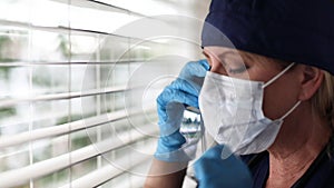 Stressed Doctor or Nurse On Break At Window Removing PPE, Face Mask and Protective Glasses