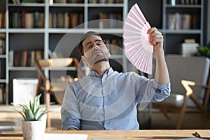 Stressed exhausted young 30s businessman using paper fan.