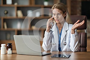 Stressed Female Doctor Talking On Cellphone And Using Laptop In Clinic