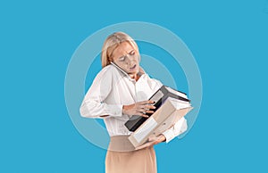 Stressed female secretary with heap of papers talking on smartphone on blue studio background