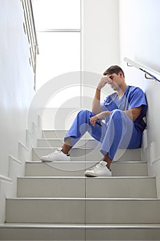 Stressed Male Doctor Or Nurse Wearing Scrubs Sitting On Stairs In Hospital 