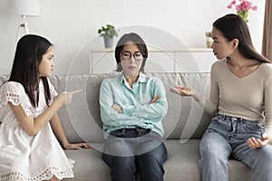 Stressed mature Asian woman sitting on couch while adult daughter and granddaughter arguing with her at home