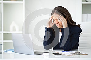 Stressed mature businesswoman sitting at workplace, suffering from headache