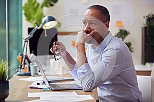 Stressed Mature Male Architect Working In Office At Desk On Plans For New Building