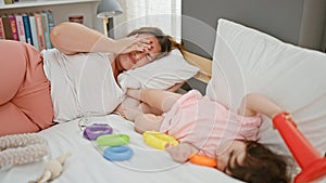 Stressed mother and daughter lying in bed as baby plays with toys in bedroom - a poignant family moment