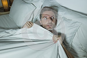 Stressed and scared man alone in bed awake at night in fear after having a nightmare feeling paranoid holding the blanket in