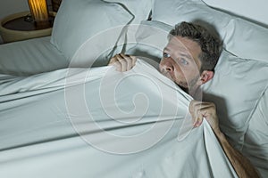Stressed and scared man alone in bed awake at night in fear after having a nightmare feeling paranoid holding the blanket in