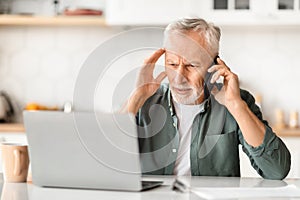 Stressed senior man using phone and laptop, sitting at desk in kitchen