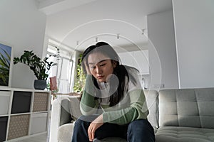 Stressed unhappy Asian woman sits on sofa at home thinking about financial problems. Heartbreak.