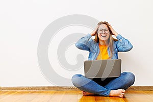 Stressed woman with laptop sitting on floor against white wall holding her head