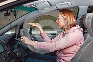 A stressed woman is sitting behind the wheel of a car and cursing, points to the road with her hand. Side view.