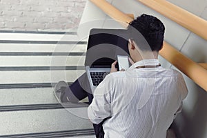 Stressed young Asian business man sitting and using mobile smart phone and laptop at staircase of office