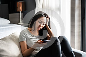 Stressed young Asian woman suffering on depression while holding mobile phone on hand sitting on couch in living room at home