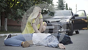 Stressed young blond woman checking pulse of body hit by car outdoors. Portrait of frustrated young Caucasian female