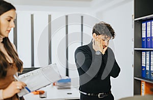 Stressed young businessman with hand on face in office, female colleague in background