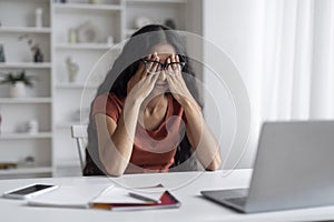 Stressed young eastern businesswoman sitting at workplace, rubbing eyes