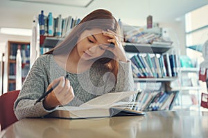 Stressed young female student sitting at wooden table and reading book during exam preparation in library