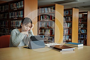 Stressed young female student sitting at wooden table and reading book during exam preparation in library