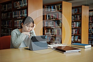 Stressed young female student sitting at wooden table and reading book during exam preparation in library