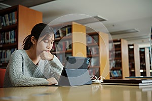 Stressed young female student sitting at wooden table and reading book during exam preparation in library