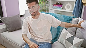 Stressed young hispanic man at home, lying on sofa, covering face with cushion, closing laptop, expressing unhappiness indoors
