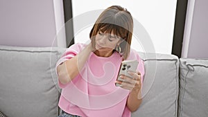 Stressed young hispanic woman suffering from neck pain while texting on her smartphone at home