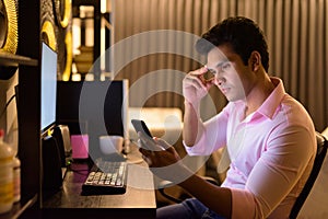 Stressed young Indian businessman using phone while working overtime at home late at night