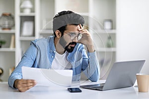 Stressed Young Indian Man Looking At Laptop Screen And Holding Documents