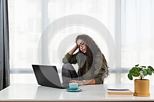 Stressed young woman holding head in hands and feeling demotivated while sitting at her home office and working remotely on laptop