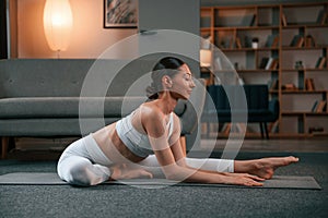 Stretching exercise. Young woman with slim body type in white sportswear is indoors doing yoga