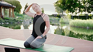 Stretching in nature. Beautiful fitness woman in sportswear sitting on yoga mat near beautiful lake and stretching neck