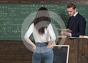 Strict professor in glasses reading a book at rostrum. Female brunette student holding hands in rear jeans pockets while