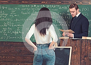 Strict professor in glasses reading a book at rostrum. Female brunette student holding hands in rear jeans pockets while