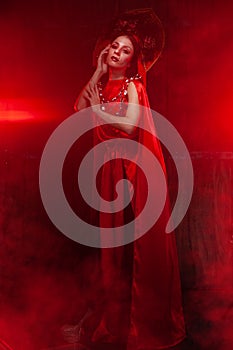 Strict stylish girl in a fashionable long red dress and a voluminous hat on the background of smoke in the Studio at