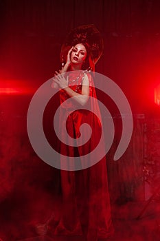 Strict stylish girl in a fashionable long red dress and a voluminous hat on the background of smoke in the Studio at