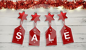 A string of christmas star decorations, lights and tinsel, with for sale written on red labels, on destressed white wood