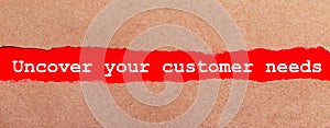 A strip of red paper under the torn brown paper. White lettering on red paper UNCOVER YOUR CUSTOMER NEEDS. View from above