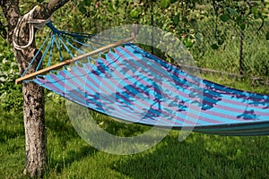Striped blue and blue fabric hammock hanging in the shade of trees in the garden on a sunny warm day.