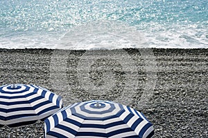 Striped blue and white umbrellas on a pebble beach on the Promenade des Anglais in Nice, France, await guests. Blue waves roll