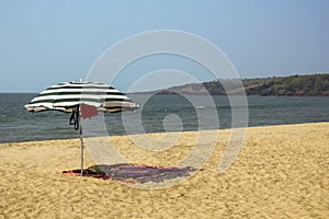 Striped large umbrella and sarong on the yellow sand of the beach against the ocean with sea waves under a clear blue sky