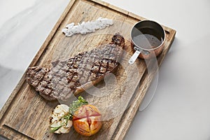 Striploin steak served with roasted vegetables and gravy sauce on cutting board, white marble background