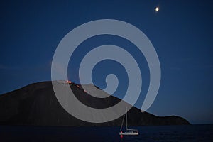 A Stromboli volcano spews lava while a sailboat sails past, with the moon above