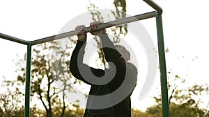 Strong athlete in black sportclothes doing pull-up on horizontal bar. Sun shines on the background and trees around him