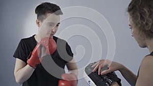 Strong athletic female in boxing gloves makes punches on paw held by woman trainer in gym. Training of professional