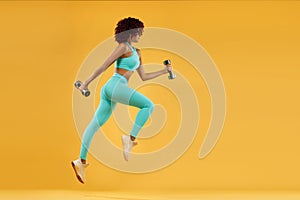 Strong athletic, woman sprinter or runner, running on yellow background with dumbbells wearing sportswear. Fitness and sport motiv