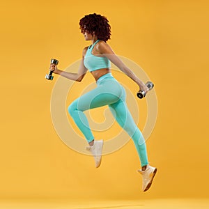 Strong athletic, woman sprinter or runner, running on yellow background with dumbbells wearing sportswear. Fitness and sport motiv