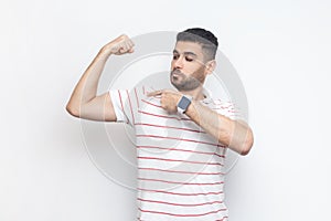 Strong attractive man in t-shirt standing with raised hand, pointing at his biceps, looks proud.