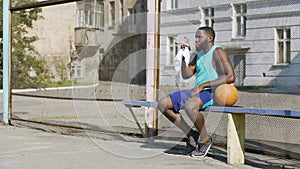 Strong basketball player sitting on bench and drinking mineral water, sport